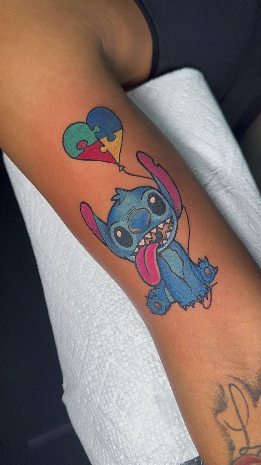 Stitch holding puzzle-piece heart balloon, forearm tattoo